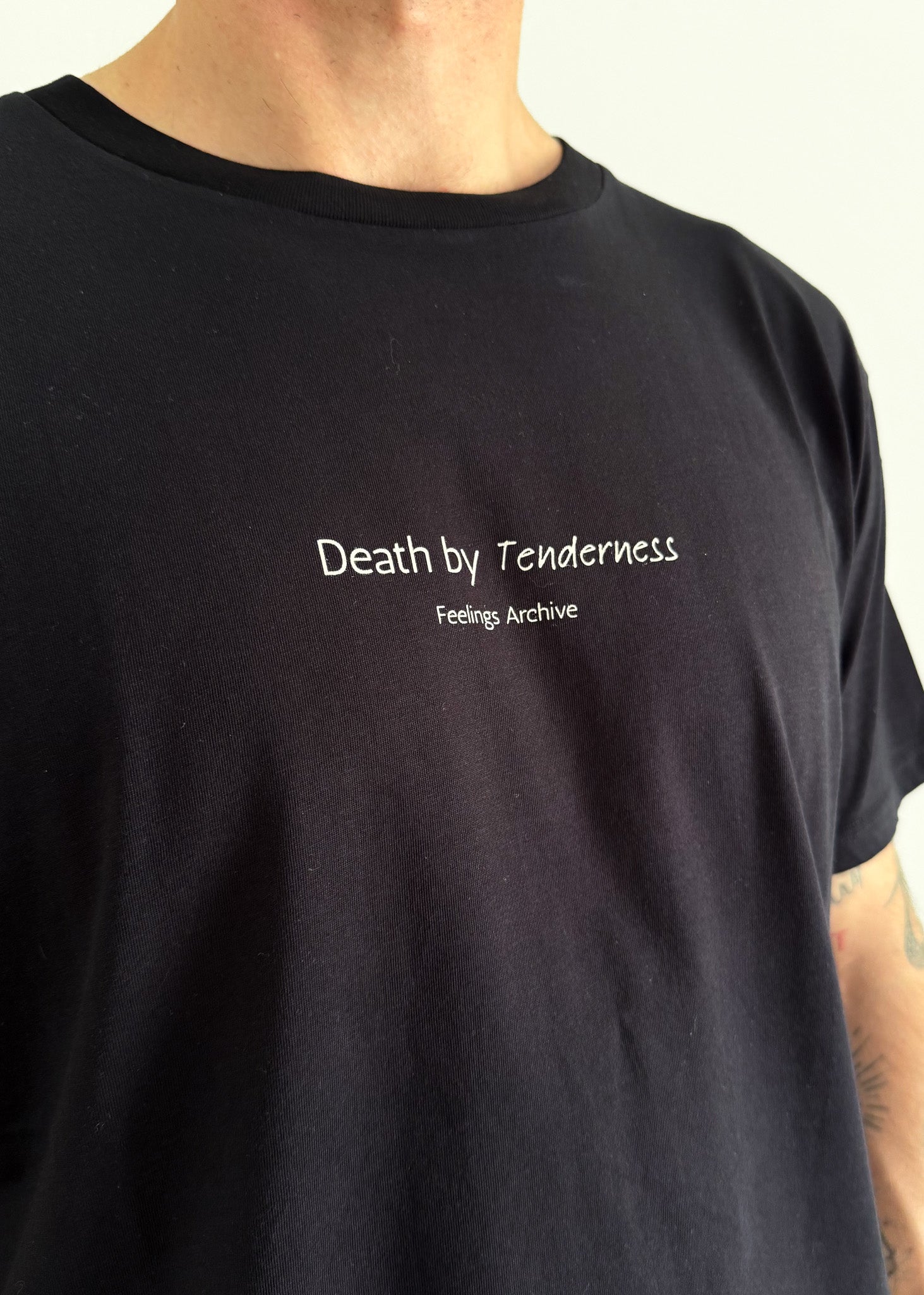 A close-up of a person wearing a black crewneck t-shirt with white text on the chest that reads 'Death by Tenderness' and 'Feelings Archive'.
